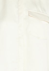 Camicia di seta color crema con taschino sul petto, texture liscia e un delicato lucido. Design classico con chiusura a bottoni e colletto.