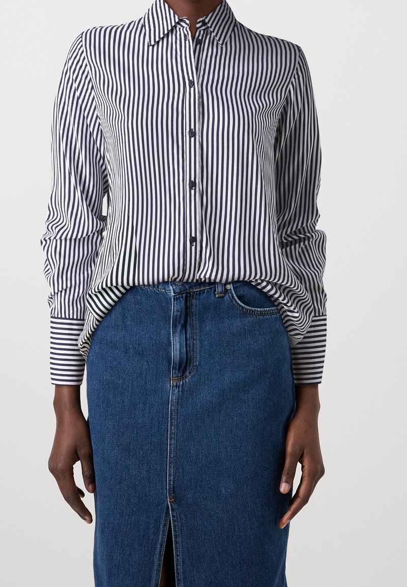 Personne portant une chemise à rayures verticales noires et blanches boutonnée et une jupe en jean bleue fendue devant, debout sur un fond uni.