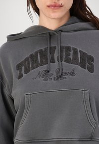 Felpa grigia con tasca a kangaroo frontale, cappuccio con cordino e logo in rilievo "TOMMY JEANS New York" ricamato in filo scuro. Tessuto morbido.