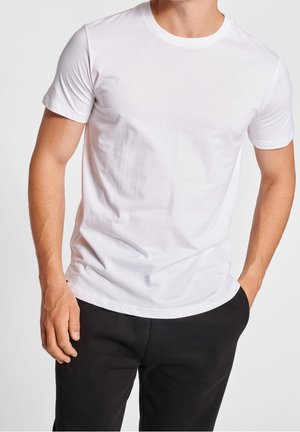 Mężczyzna w gładkiej białej koszulce z krótkim rękawem i czarnych spodniach, z prawą ręką dotykającą spodni i lewą ręką w kieszeni.