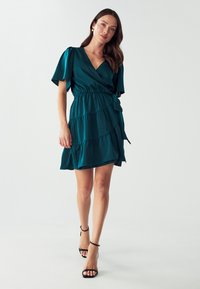 Teal Wickelkleid aus glattem Stoff mit kurzen, flatternden Ärmeln, mehrlagigem Rüschen-Saum und Taillengürtel. Kombiniert mit schwarzen Riemchen-Heels.