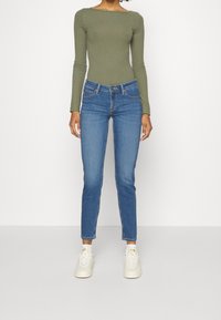 Olivgrön ribbad långärmad bodysuit, kombinerad med medelblå skinny jeans med knappstängning fram och vita sneakers.