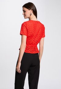 Haut en dentelle rouge avec des bords festonnés, des manches courtes et une taille ajustée, associé à un pantalon noir. Motif floral détaillé sur tout le tissu.