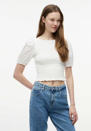 Ung kvinde iført en hvid, puffet, smocked crop top og højttaljede blå denim jeans mod en ensfarvet baggrund.