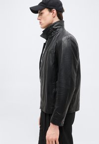 Άνδρας με μαύρο δερμάτινο μπουφάν με ψηλό γιακά, μαύρο καπέλο baseball και σκοτεινό παντελόνι με λεπτές λωρίδες, στέκεται προφίλ μπροστά σε απλό φόντο.