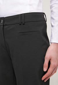 Pantalons noirs ajustés avec une texture lisse, dotés d'un devant plat et d'une poche latérale, associés à une chemise blanche à manches longues.