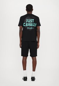 Crna pamučna majica s plavim grafičkim natpisom "JUST CAVALLI MILANO" na leđima, uparena s crnim šorcem i crnim cipelama s bijelim čarapama.