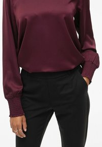 Bordeaux satin bluse med lange ærmer, med detaljer i smock ved manchetten. Kombineret med tætsiddende sorte bukser. Glat tekstur og elegant design.