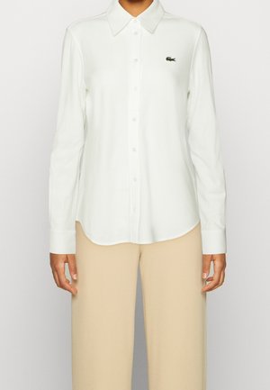 Personne portant une chemise blanche à manches longues avec col et pantalon beige, montrant le haut du corps et les bras de face.