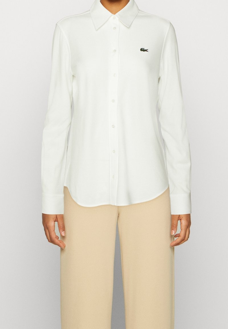 Personne portant une chemise blanche à manches longues avec col et pantalon beige, montrant le haut du corps et les bras de face.