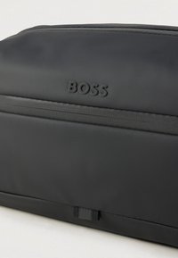 Melnā sintētiskā soma ar gludu tekstūru, reljefā iespiestu "BOSS" logotipu un horizontālu rāvējslēdzēja nodalījumu augšpusē.