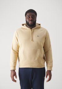 Sudadera beige con un bolsillo frontal, capucha con cordones y pequeño logo verde. Combinada con pantalones azul oscuro, de corte relajado y textura suave.