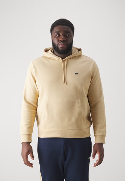 Sweat à capuche beige avec une poche avant, un cordon de serrage à la capuche et un petit logo vert. Associé à un pantalon bleu marine, au fit décontracté et à la texture douce.