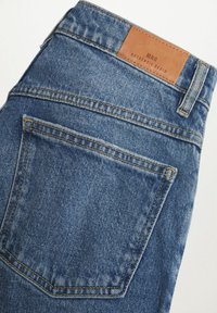 Jean en denim bleu plié montrant la poche arrière et un patch de marque en cuir marron clair avec l'inscription "MNG Authentic Denim" sur fond blanc.