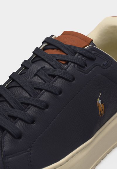 Baskets en cuir bleu marine avec lacets bleu marine, étiquette de languette en cuir beige, semelle crème et logo de joueur de polo brodé sur le côté.