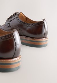Scarpe eleganti in pelle marrone con dettagli brogue, punta arrotondata e tacco impilato in legno con accenti colorati.