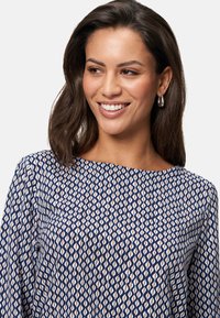Navy blouse met geometrisch crèmekleurig en bruin patroon, ronde halslijn, wijdvallende mouwen en een zachte stoftextuur. Zilveren oorbellen in de vorm van een ring.