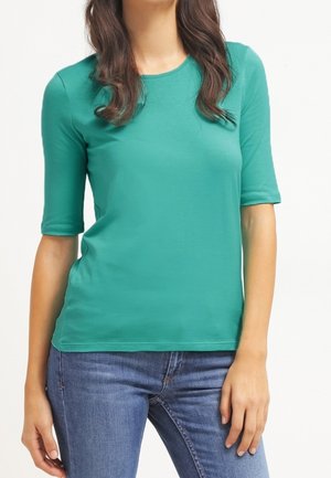 Vrouw draagt een aansluitend teal kortemouwen T-shirt en blauwe jeans, staand tegen een effen witte achtergrond.