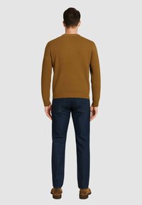 Maglione marrone con texture e collo a costine, abbinato a jeans in denim blu scuro. Modello mostrato di spalle, in piedi su uno sfondo grigio chiaro.