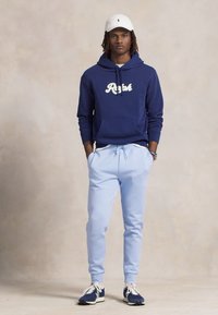 Sweatshirt azul-marinho com o texto "Ralph", calças de fato azul-claro, boné branco e ténis azul-marinho. Tecido casual, ajuste descontraído e detalhes com cordão.
