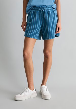 Osoba ubrana w niebieskie i granatowe szorty w paski ze sznurkiem, jasnoniebieską bluzkę i białe trampki, stojąca na jednolitym szarym tle.