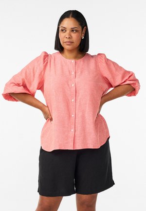 Kvinde med skulderlangt sort hår iført en lyserød knap-op-bluse med puffede ærmer og sorte shorts til knæet, stående med hænderne på hofterne.