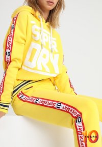 Gele hoodie en bijpassende joggingsbroek met groot wit "SUPERDRY" logo. Voorzien van rode accentstroken met tekst langs de mouwen en benen.