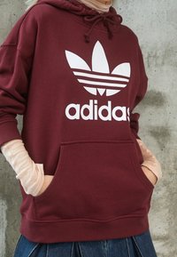 Felpa con cappuccio bordeaux con logo adidas bianco e tre strisce. Presenta una tasca anteriore e un tessuto morbido con una vestibilità comoda.