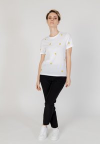 Witte T-shirt met korte mouwen, voorzien van kleine gele en groene citroenborduursel, gecombineerd met zwarte broek en witte sportschoenen.