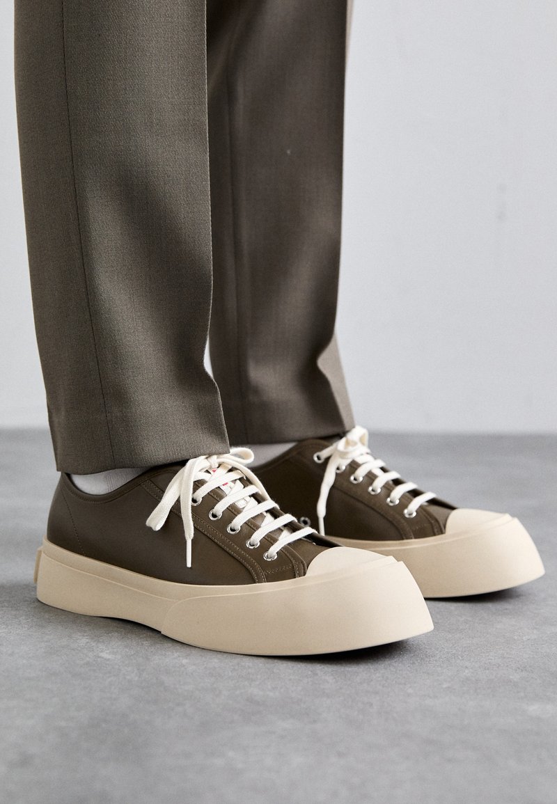 Baskets basses vert olive avec des semelles épaisses beige et des lacets blancs, portées avec un pantalon marron sur un sol gris.