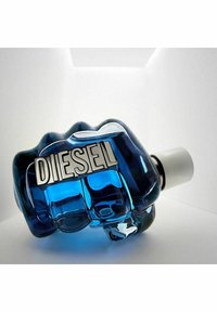 Sininen lasipullo, joka on muotoiltu puristetun nyrkin näköiseksi. Pullossa on hopeinen "DIESEL"-etiketti ja valkoinen korkki. Pullon pinta on sileä ja valon heijastukset näkyvät sen yksityiskohdissa.