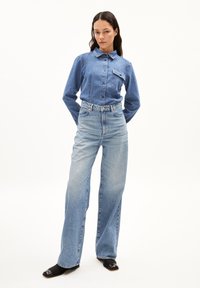 Blaue Jeanshemd mit Kragen und einer Brusttasche, kombiniert mit hoch-taillierten hellblauen Jeans. Das Model trägt schwarze High Heels.