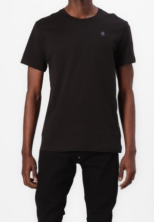 Personne portant un t-shirt noir uni à manches courtes et un pantalon noir, debout devant un fond blanc, torse et bras visibles.