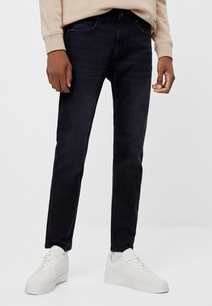 Jeans in denim scuro con una vestibilità slim, caratterizzati da un classico design a cinque tasche. Indossati con sneakers bianche e una felpa beige.