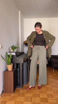 Femme ajuste un blazer vert olive tout en portant un pantalon ample vert clair, un haut noir et des chaussures pointues marron dans un salon confortable.