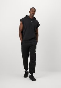 Man in een zwarte mouwloze hoodie en zwarte joggers met witte tekst, staand met zijn handen in zijn zakken tegen een effen lichte achtergrond.
