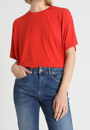 Rotes Kurzarmtop aus glattem Stoff, kombiniert mit hellblauen Denim-Jeans mit Knopfverschluss und Fronttaschen.