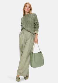 Grüner Kabelstrickpullover, hellgraue plissierte Hose und eine große grüne Lederhandtasche. Die Schuhe haben eine weiche Textur mit Verzierungen.