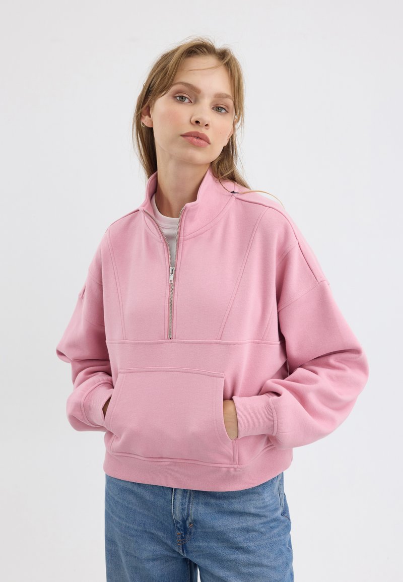 Roze zip-up sweatervest met grote voorkauw, ribbelmanchetten en een ontspannen pasvorm. Heeft een staande kraag en naaddetails. Blauwe jeans gedragen.
