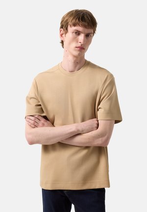 Ung mand med kort brunt hår iført en beige kortærmet skjorte og mørke bukser, står med armene over kors mod en hvid baggrund.