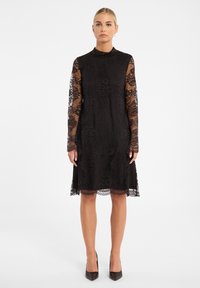 Zwarte kanten jurk met een hoge halslijn, lange doorschijnende mouwen en een geschulpte zoom. Bloemenpatronen overal in de jurk versterken de getextureerde uitstraling.