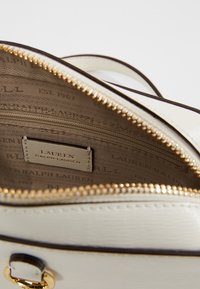 Borsa in pelle bianca con zipper dorata parzialmente aperta, che mostra l'interno in tessuto beige stampato con "Lauren Ralph Lauren" e un'etichetta del marchio cucita.