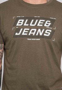T-shirt en coton vert olive avec un texte graphique audacieux en blanc "BLUE&JEANS" et des rayures, aux détails de marque dans un design moderne.