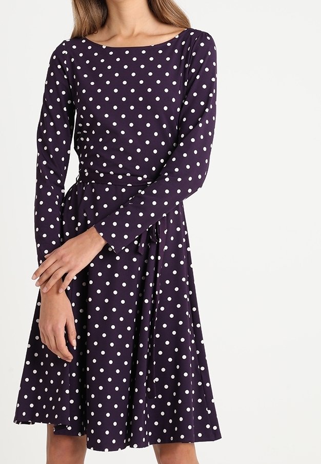 Kvinna i en knälång marinblå klänning med vita prickar, långa ärmar och ett knutet midjebälte, stående mot en vit bakgrund.