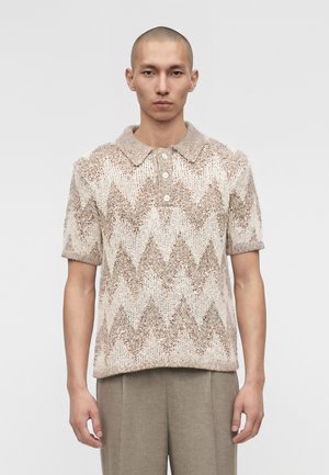 Νέος άνδρας φορώντας ένα μπεζ και ανοιχτό καφέ πλεκτό polo με κοντά μανίκια και σχέδιο ζικ-ζακ, και παντελόνι σε απόχρωση ταουπέ, στέκεται μπροστά σε ένα απλό φόντο.