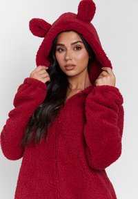Túnica roja y esponjosa con capucha que tiene orejas de oso. Presenta una cremallera, grandes puños y una textura suave para mantener el calor.