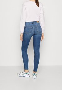Jeans de mezclilla azules de talle alto con corte slim, bolsillos traseros y costuras en contraste, combinados con zapatillas blancas y verdes.