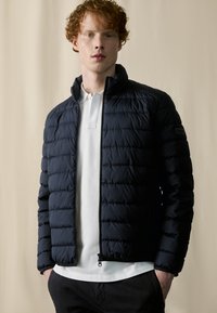 Navy puffer jacket with a quilted design, high collar, and zip closure. Worn over a white polo, featuring side pockets and textured fabric.