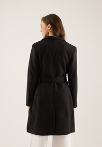 Vrouw met lang bruin haar, gekleed in een zwarte mantel met riem en witte broek, met de rug naar de camera tegen een effen lichte achtergrond.