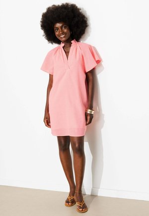 Femme avec coiffure afro souriante, portant une robe courte rose avec des manches volantées et des sandales marron, debout contre un mur blanc.